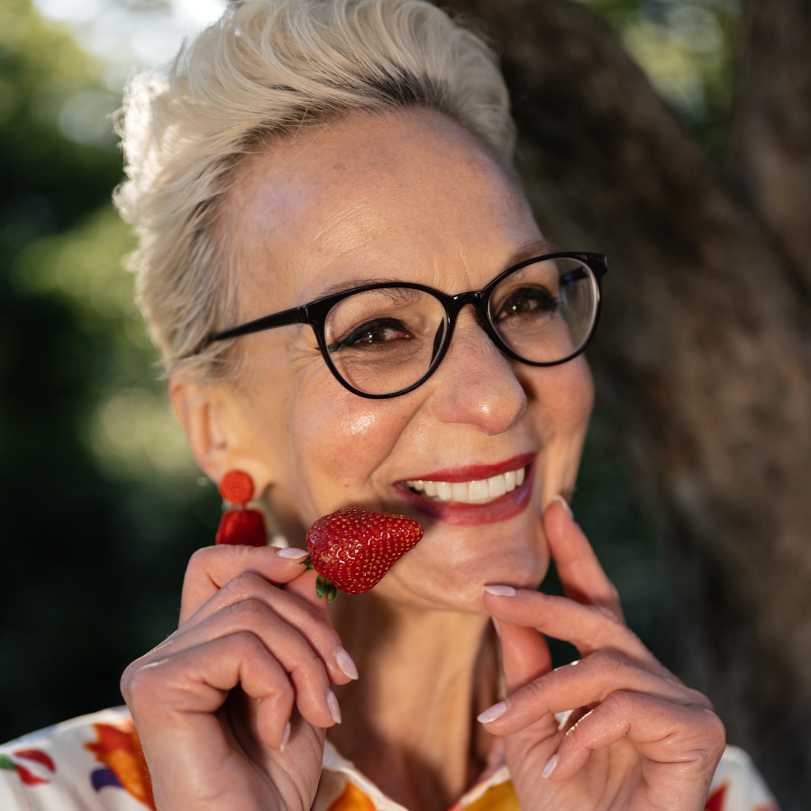 Donna over 50 sorridente con trucco perfetto, tenendo una fragola vicino alle labbra.