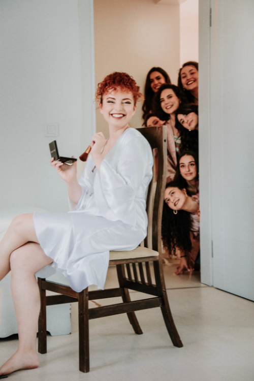 Sposa in attesa di essere truccata con amiche storiche sullo sfondo.